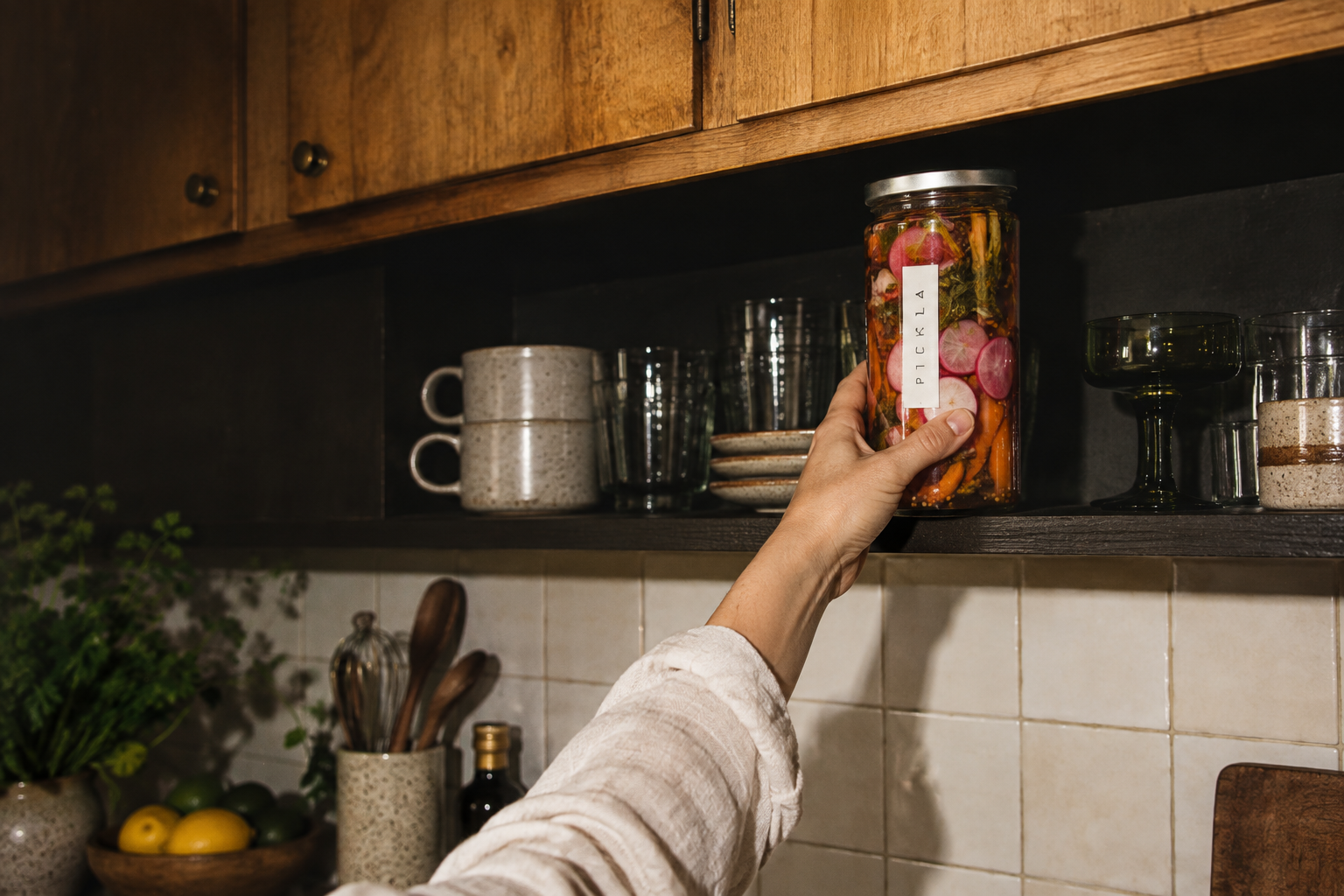 A hand placing a jar of pickles on a warm wooden kitchen shelf